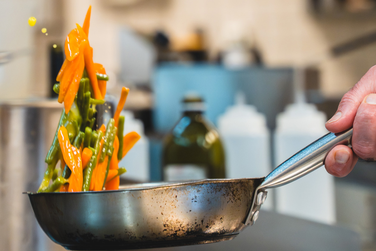Koch schwenkt frisches Gemüse – Karotten und grüne Bohnen – in einer Edelstahlpfanne in der Küche des Bella Vista