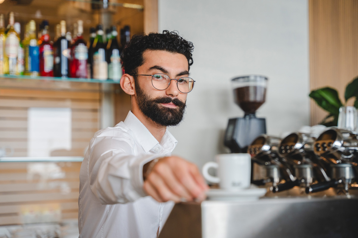 Junger Barista mit Brille serviert einen Espresso an der professionellen Kaffeemaschine im Bella Vista